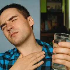 Mężczyzna z zamkniętymi oczami trzyma szklankę wody i dotyka gardła.
