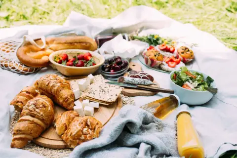 Piknikowy stół z croissantami, owocami, wędliną, sałatką i napojami.
