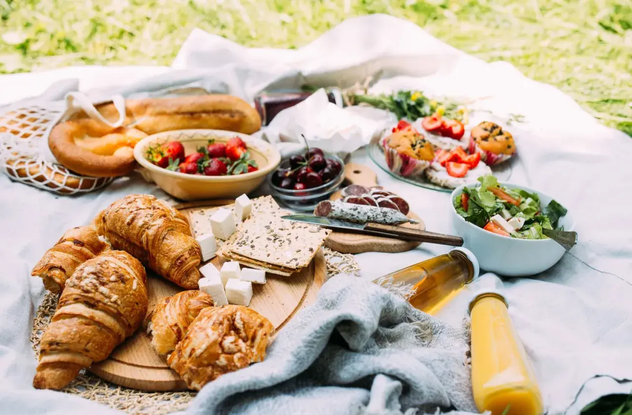 Piknikowy stół z croissantami, owocami, wędliną, sałatką i napojami.