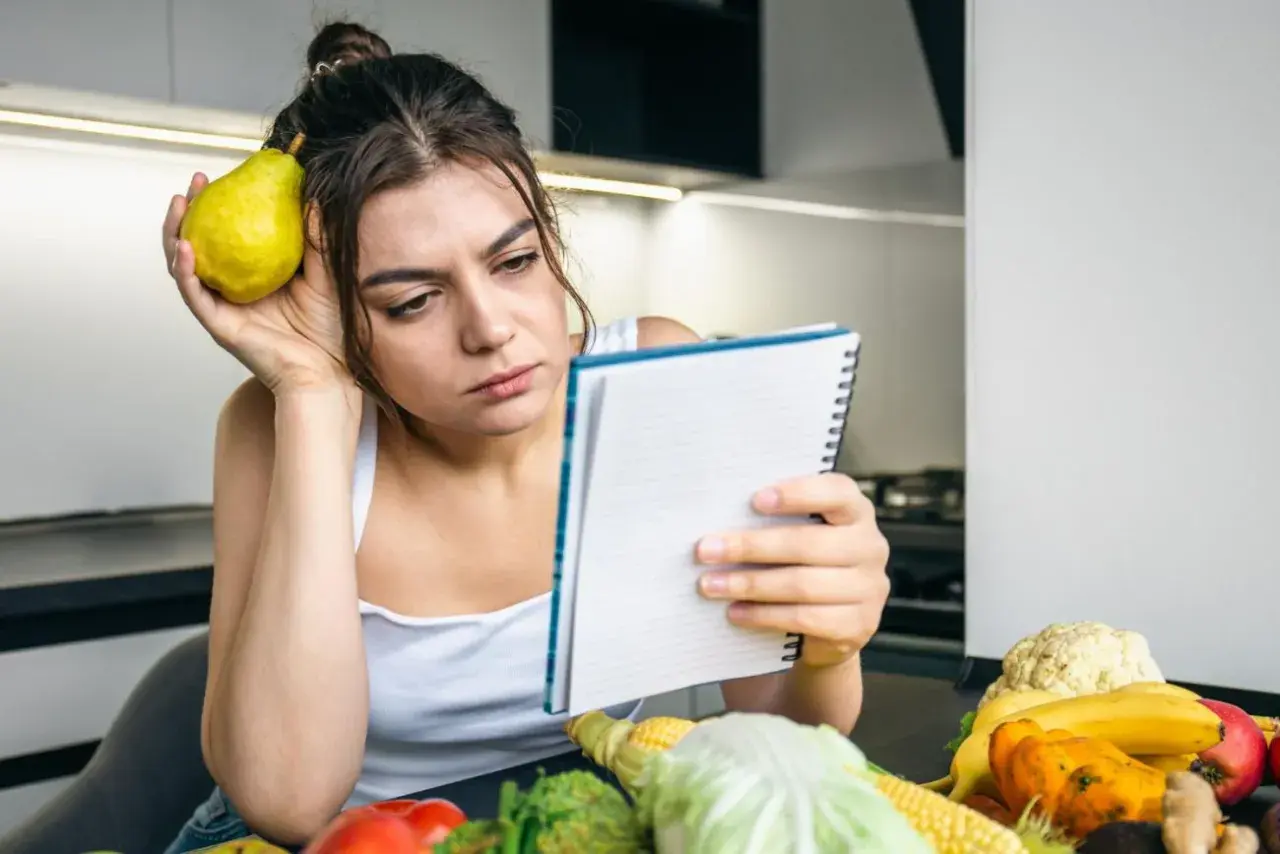 Zamyślona kobieta z gruszką przy uchu przegląda notatnik. Na stole warzywa i owoce, jakby planowała dietę 500 kcal.